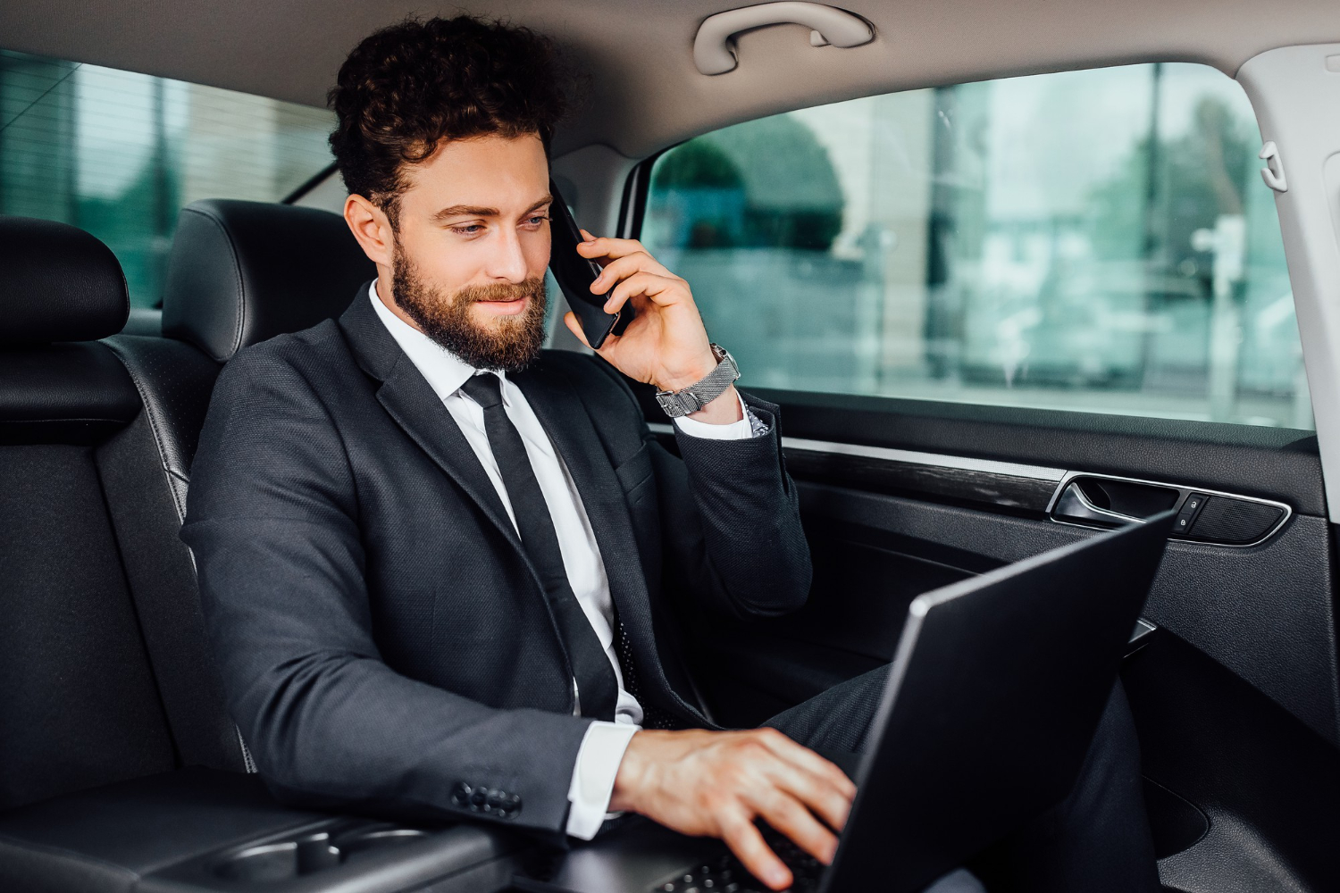 handsome bearded smiling businessman working his laptopand speaking mobile phone backseat car